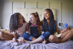 Three young girls sit cross-legged on a bed, sharing a joyful moment. 